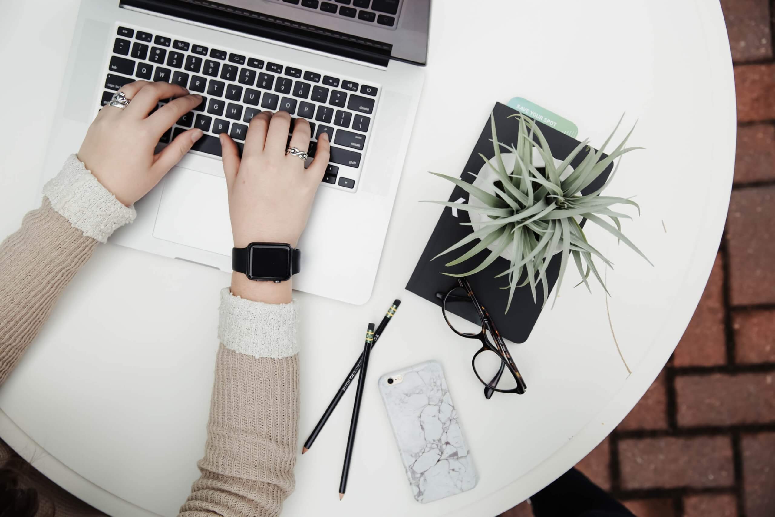 Vrouw schrijft op een laptop en naast haar staat een plant en liggen wat spullen op tafel